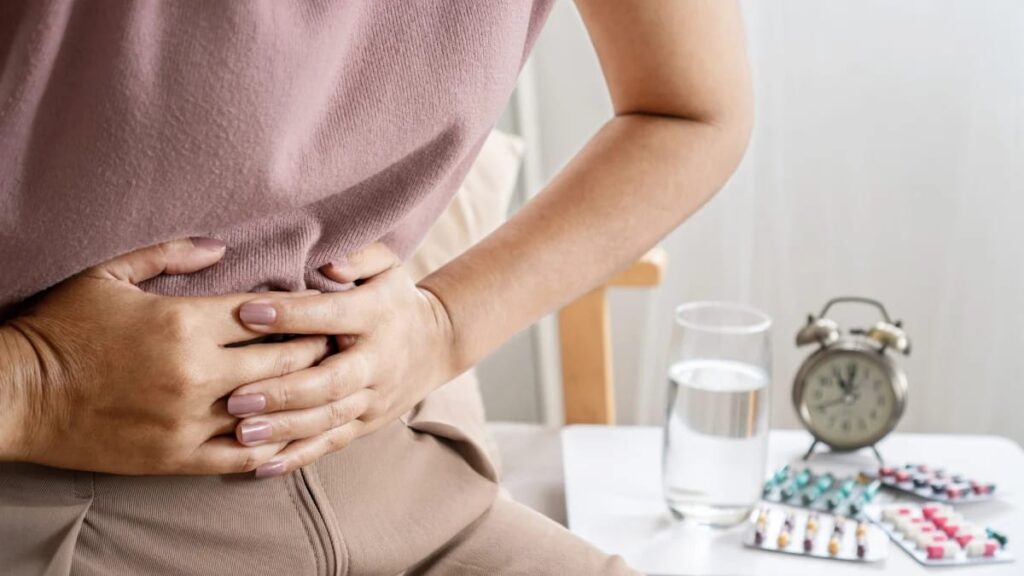 Inquadratura ravvicinata di una persona che preme con forza le mani sull'addome in segno di dolore e disagio. Sullo sfondo, un comodino con un bicchiere d'acqua, diverse confezioni di medicinali in blister e una sveglia, elementi che suggeriscono la gestione cronica dei sintomi del colon irritabile.